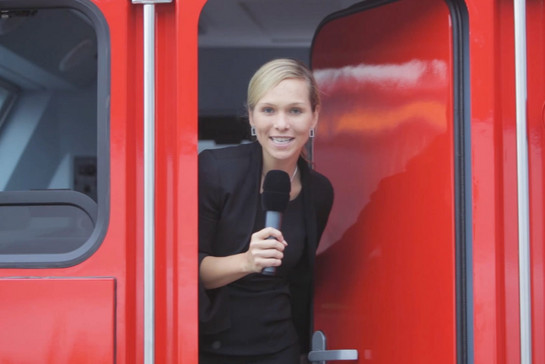 Junge Frau steht in einem roten Zug in der Tür, sie hält ein Mikrofon in der Hand. 