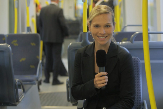 Eine junge Frau mit Mikrofon sitzt in einem Bus, hinter ihr sind zwei Personen zu sehen. 