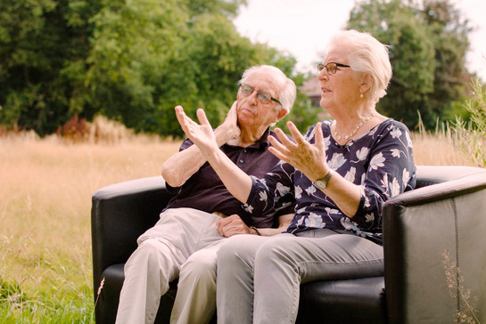 Standbild von zwei älteren Personen die auf einem schwarzen Sofa sitzen
