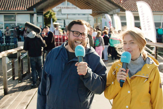 Zwei Journalisten stehen mit Mikrofonen an einem Steg