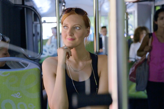 Eine Frau sitzt in einem Stadtbus, im Hintergrund sind weitere Fahrgäste. 