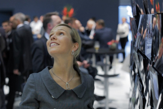 Eine junge Frau, sie lächelt. Im Hintergrund sind Menschen in Business-Kleidung zu sehen. 