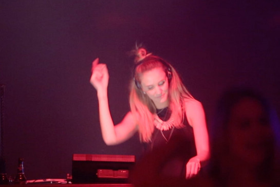 Eine Frau an einem DJ-Pult, sie hat Kopfhörer auf und bewegt sich zur Musik. 