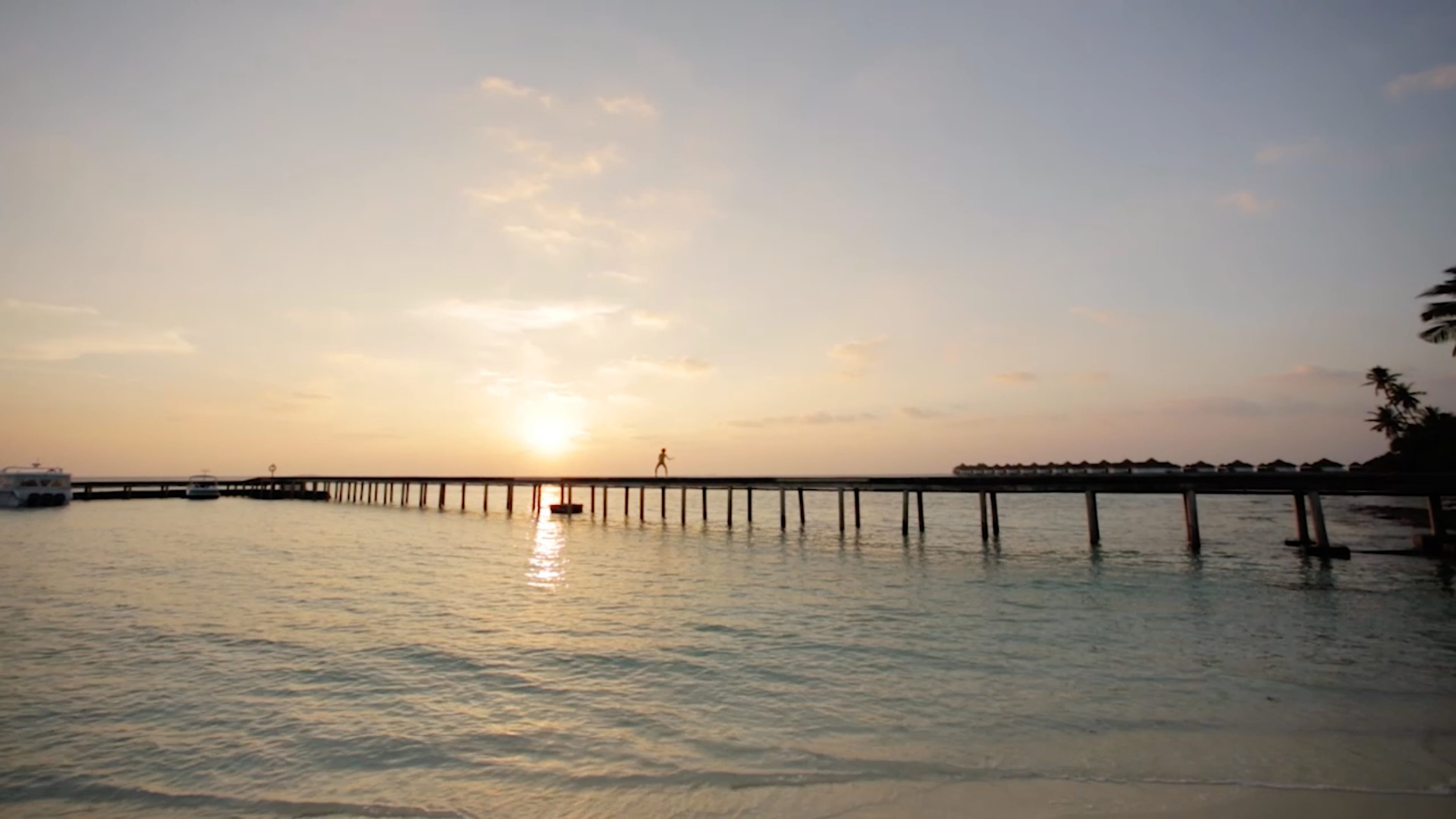 Ein langer Steg auf dem Wasser, darauf eine Person. Dahinter Sonnenuntergang. Am Strand scheinen Palmen zu sein. 