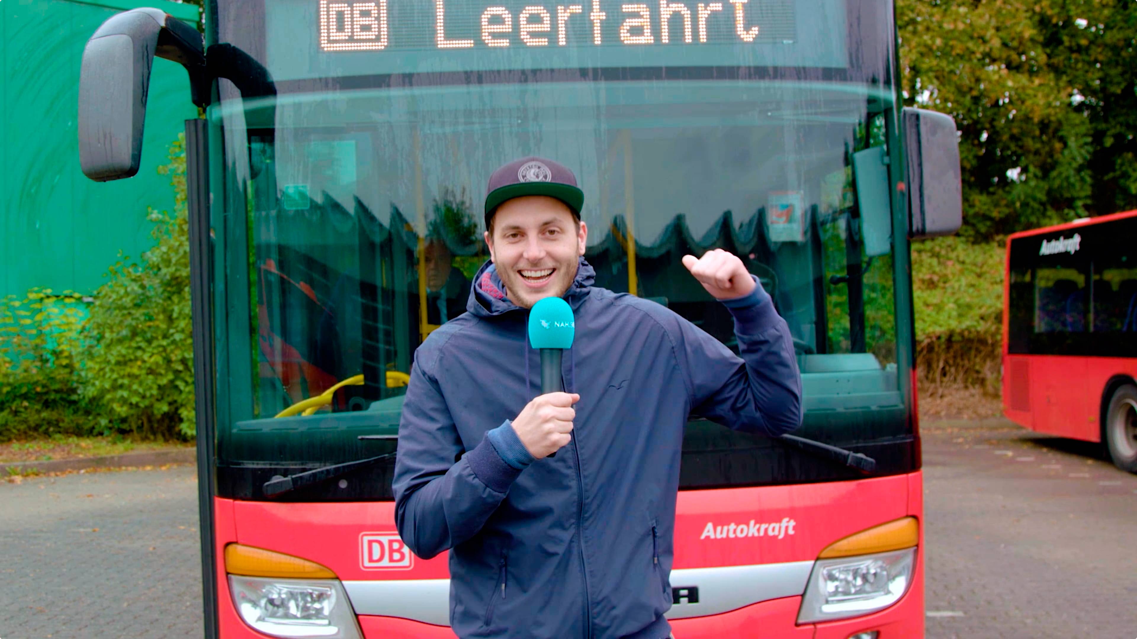 Ein Mann mit Mikrofon zeigt auf einen Bus hinter sich