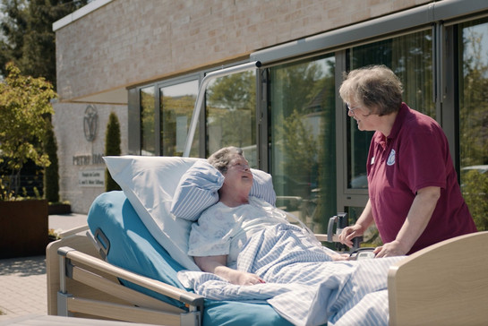 Eine kranke Frau liegt vor dem Hospiz in ihrem Bett und wird von einer Pflegerin betreut.