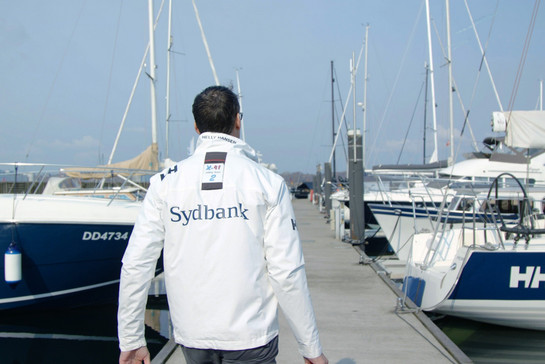 Testimonial Sven Christensen läuft auf einem Steg Richtung Segelboot und trägt eine weiße Jacke, mit dem Schriftzug Sydbank auf dem Rücken.