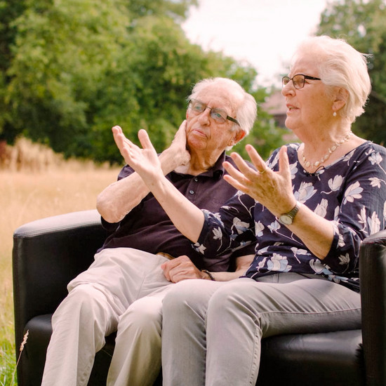 Standbild von zwei älteren Personen die auf einem schwarzen Sofa sitzen
