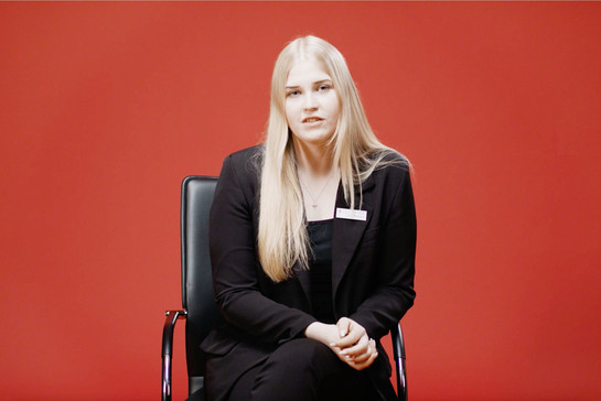 Eine junge Frau sitzt vor einer roten Wand auf einem Bürostuhl und schaut mit gefalteten Händen in die Kamera. Sie trägt ein Namensschild von der Sparkasse. 