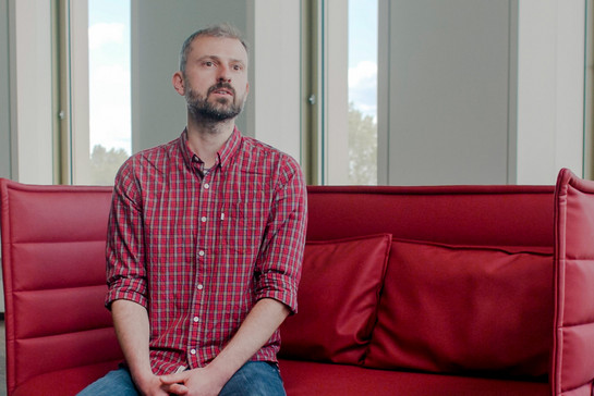 Ein Mann in einem karierten Hemd und Jeans sitzt auf einem roten, modernen Sofa. 