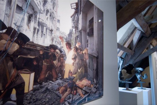 Das Kunstwerk zeigt einen Kiregsschauplatz: Eine Menschengruppe steht dicht zusammengedrängt vor zerstörten Häuserfassaden. Gegenüber stehen vermummte Menschen und richten Waffen auf die Gruppe.