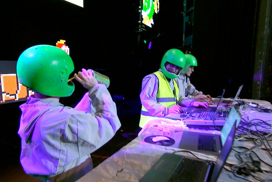 Drei Männer mit grünem Helm stehen an einem DJ Pult
