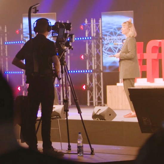 Auf einer kleinen Bühne steht eine Moderatorin, ein Kameramann  filmt sie. 