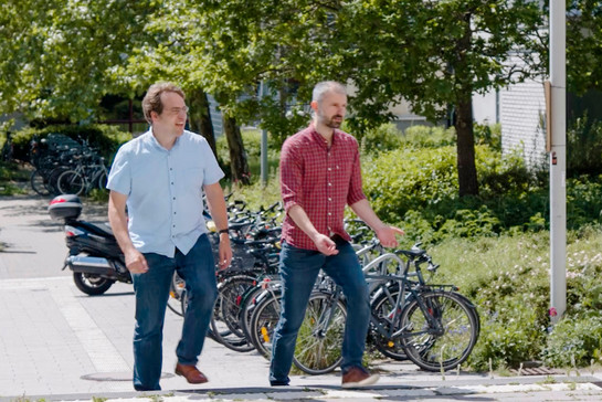 Zwei Männer in Jeans und Hemd auf einem Bürgersteig, im Hintergrund parkende Fahrräder. 