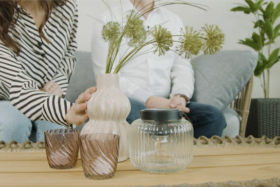 Vase mit Blume, 2 Teelichtgläser und eine Glaslaterne stehen auf einem Tisch, im Hintergrund eine Frau und ein Mann auf einem Sofa