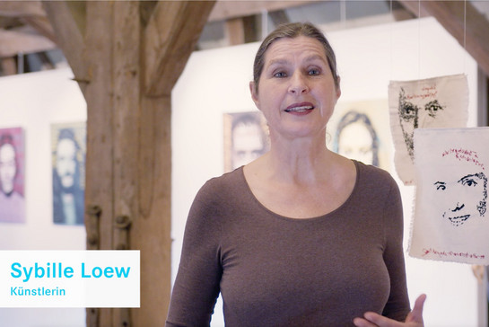 Die Künstlerin Sybille Loew spricht im Museum über ihre Kunst. Neben und hinter ihr hängen Stoffquadrate mit Skizzen von unterschiedlichen Gesichtern von der Decke.