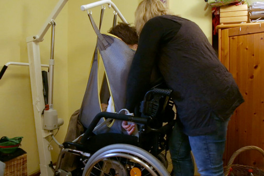 Eine Frau wird mit einer Trage in einen Rollstuhl gesetzt