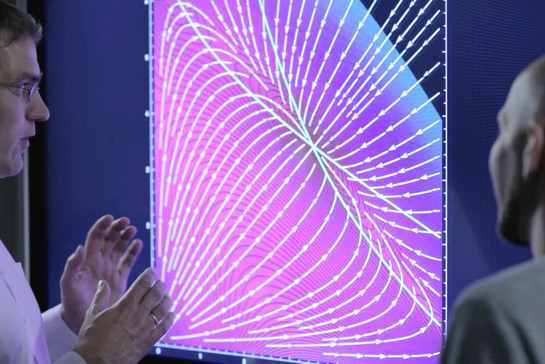 Zwei Männer schauen auf einen großen Bildschirm, darauf eine Grafik mit vielen Pfeilen. 