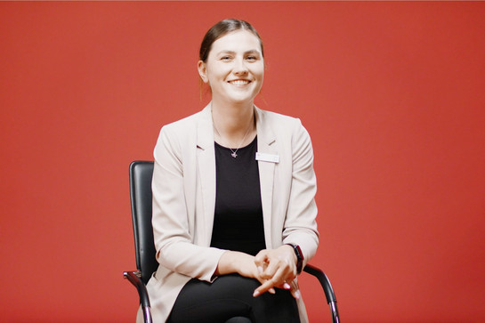 bb_nospa_azubi_teaser_06.jpg Junge Frau mit braunen Haaren sitzt vor einer roten Wand auf einem Bürostuhl