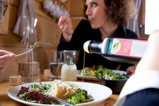 bb_cellagon_teaser_02.jpg Zwei Personen sitzen an einem Tisch, sie essen Salat mit Ofenkartoffeln. Jemand tut etwas Speiseöl auf den Salat, auf der Flasche steht „Cellagon“.