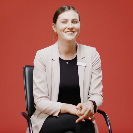 Eine junge Frau sitzt vor einer roten Wand auf einem Bürostuhl und lächelt in die Kamera. Sie trägt ein Namensschild von der Kreissparkasse am Kragen.