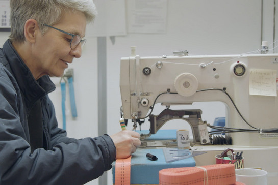 Eine Frau mit kurzen Haaren und Brille arbeitet an einer großen Nähmaschine. 