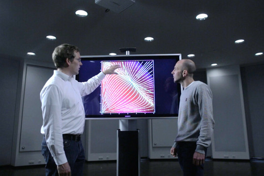 Zwei Männer in einem relativ dunklen Raum, sie schauen auf einen großen Bildschirm auf der eine Grafik gezeigt wird. 