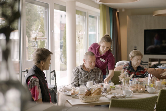 Drei ältere Menschen sitzen am Frühstückstisch und werden von einer Pflegerin betreut.