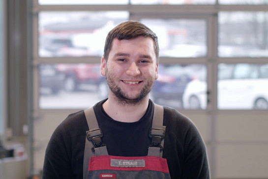 Ein KFZ Mechaniker in Arbeitskleidung lächelt in die Kamera