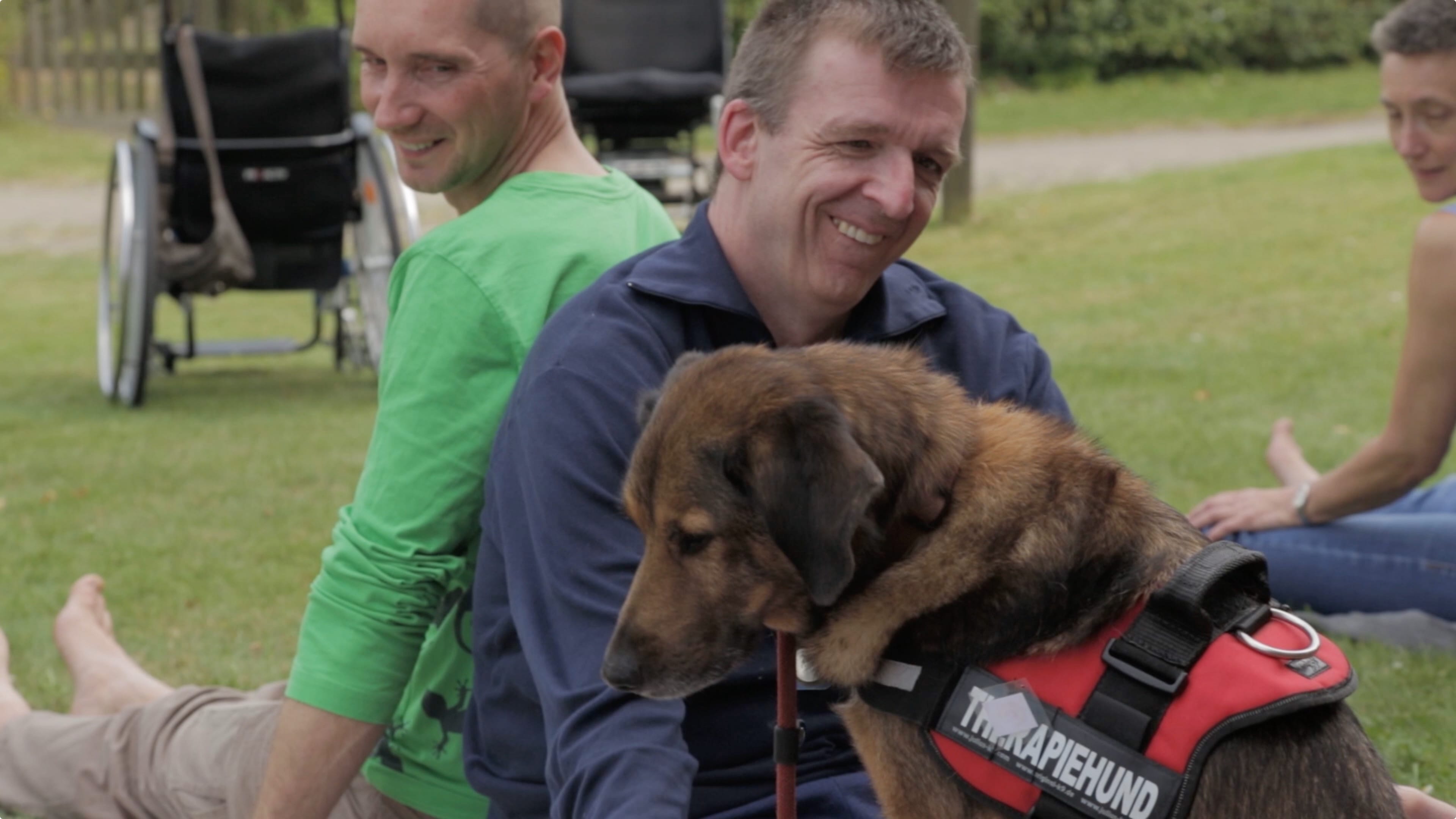 Zwei Männer sitzen mit einem Therapiehund auf dem Rasen